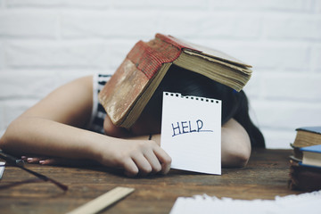 Elementary aged child sleeping at desk with book over head and holding written sign that says Help