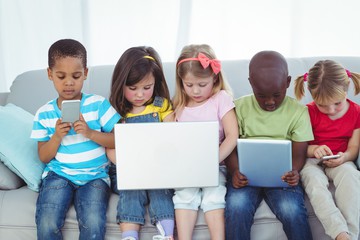 5 children sitting on sofa with various technological items like iPads, mobile phones, and a laptop computer.