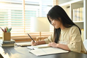 Image of smiling asian teenage student, doing homework, writing task, taking notes on desk at home